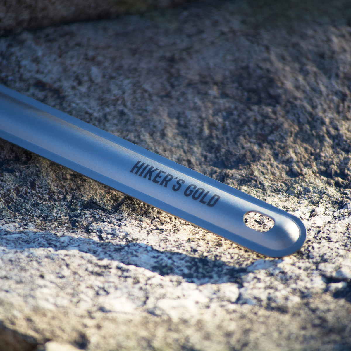 A close-up of a titanium spork handle with the engraved text 'HIKER'S GOLD' resting on a textured gray rock surface.