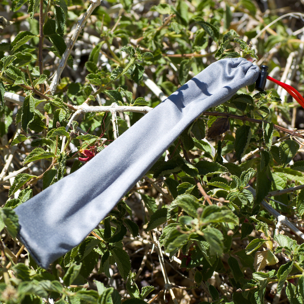 A light gray fabric pouch or sleeve with a black snap closure and a red loop, photographed hanging among green leaves and branches outdoors.