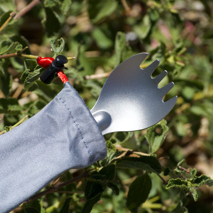 A titanium spork with a gray silicone sleeve and a red-and-black looped handle, photographed outdoors among green leaves in natural sunlight.